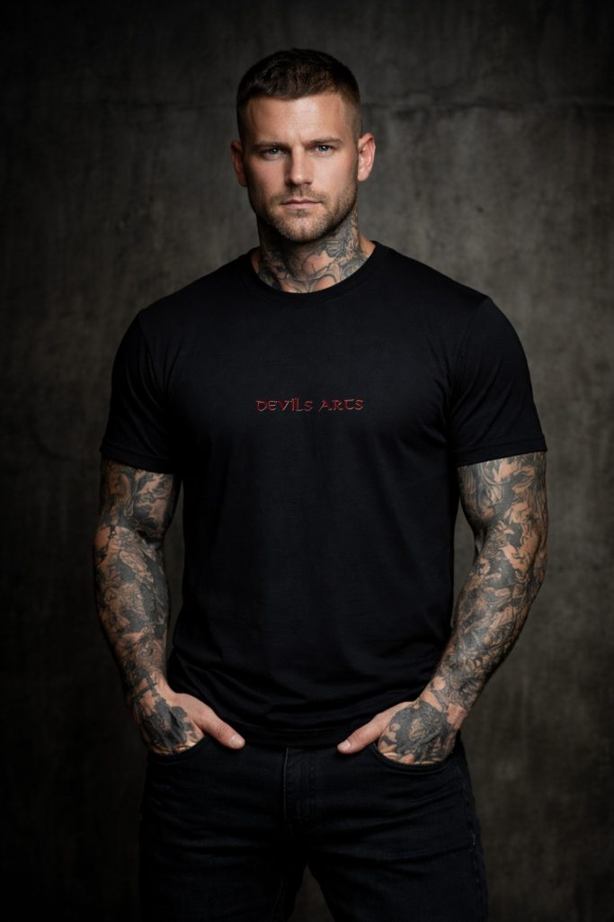 DEVILS-ARTS | WEAR – tätowiertes männliches Model mit schwarzem T-Shirt und kleiner Brustsignatur vor dunklem Hintergrund