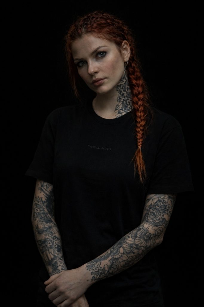 DEVILS-ARTS | WEAR – tätowiertes weibliches Model mit schwarzem T-Shirt und kleiner Brustsignatur vor dunklem Hintergrund