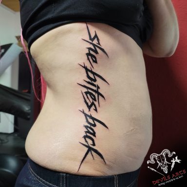 Schriftzug-Tattoo „She bites back“ in aggressiver, geritzter Optik auf der Körperseite.